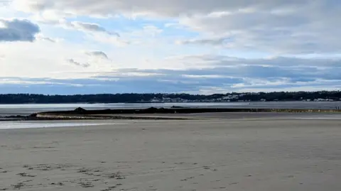 A wide shot of the Victorian Marine Lake with the sea around it, on the other side of a wall. There is a large beach, and the lake is still. There is a blue sky above with clouds.