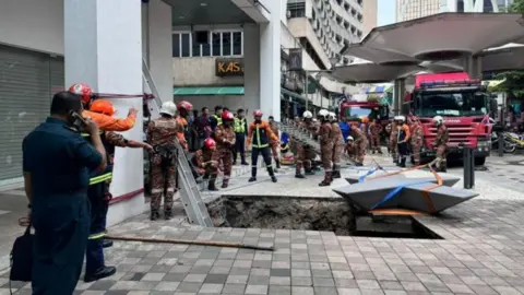 Departamento de Bombeiros e Resgate de Kuala Lumpur Equipes de resgate da Malásia tentam entrar em um buraco para salvar uma mulher que foi repentinamente engolida por uma calçada em Kuala Lumpur.