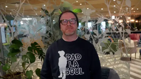Tim Everest/BBC A man wearing a beanie and glasses stood indoors in a lobby area. he is wearing a sweatshirt that reads 'Dad La Soul' and has the silhouette of a child. 