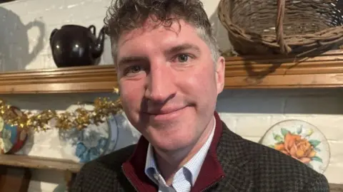 Edward Morello, MP for West Dorset, pictured from the chest up. He has short brown curly hair that is greying on the sides. He is wearing a pale shirt, a burgundy, zip-up jumper and a brown, chequered suit jacket. He is smiling at the camera. Behind him is a wooden Welsh dresser with various items on the shelves, including a woven basket.