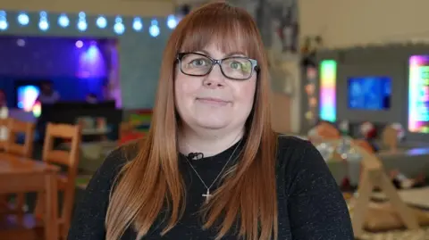 A head-and-shoulders picture of Lynn Hammel-Randall. She has long brown-red hair and a fringe. She is wearing a black jumper, glasses with a dark rim and a necklace with a cross pendant. She is sat in the cafe and the background is blurred.