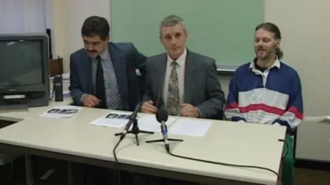 A picture of police officers Graham Sunderland and Tony Whittle, both wearing suits and sitting behind a table at a police press conference in 1994. Alongside them is Geri Gordon wearing a blue, white and red striped jumper.