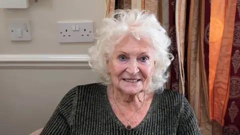 Jackie Holden sat in a chair wearing a grey jumper and a pendant