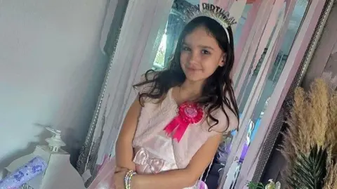 A young girl with long dark-brown hair is smiling at a camera wearing a light-pink princess dress and jewellery. She is wearing a crown which says 'Happy Birthday'.