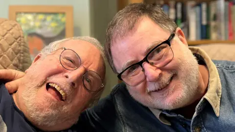 Bernard and Danny Mendoza, both smiling at the camera in a care home. Danny has his arm around his father.