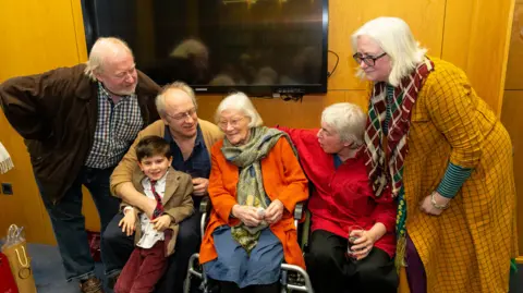 Patrick Smyth Shows Jennifer Johnston in a bright orange top with a green scarf sitting in a wheelchair with two men and a boy to the left and to women - one in a red top and the other in an orange top to the right. 