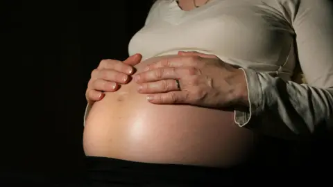 A pregnant person holding both their hands across their tummy. They are wearing a pale coloured top which is pulled up to show the bump.