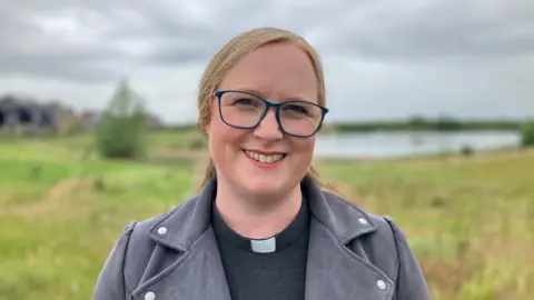 JOANNA TAYLOR/BBC Reverend Beth Cope smiling at the camera. She is wearing a dog collar, glasses grey jacket. Behind him is a lake surrounded by grass, out of focus. 