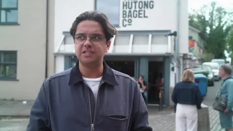 George Harman, wearing a blue jacket and white top and glasses. He is looking to the left of the camera. Behind him is a white building that says "Hutong Bagel Company". There are people behind him. 