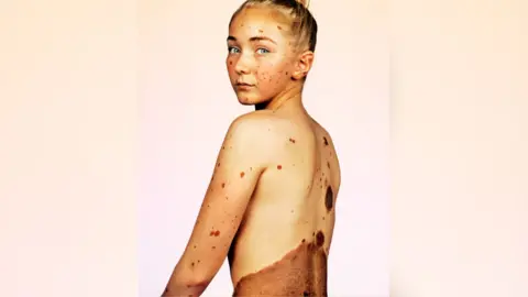 @mrelbank @caringmattersnow Clarke with her left side and back to the camera, showing the CMN on her back and CMN planets across her skin. She has her blonde hair up in a high bun and is looking over her left shoulder towards the camera. 
