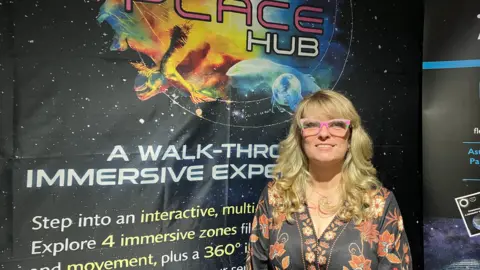 Amy Holmes/BBC A woman with long blonde, wavy hair wearing a patterned dress, standing indoors against a vibrant banner advertising an interactive, immersive experience.
