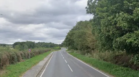 A general view of a road leading to Halten Holegate, near Spilsby. There are trees on one side of a road and hedging on the other. There are white markings on the road surface.