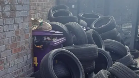 Local Democracy Reporting Service Dozens of black vehicle tyres piled beside metal alley gates between brick terraced houses.