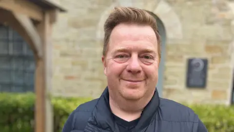 BBC Close up of Marc Neeld. The health practitioner is smiling at the camera and is wearing a black gilet. He is standing in front of a stone wall.