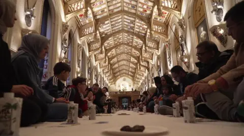 People are sat in rows  in St George's Hall facing a white table cloth. On the table cloth are plates of dates and drinks in cartons.