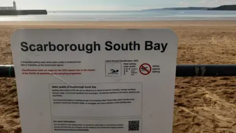 Anttoni James Numminen/LDRS A sign affixed to a metal barrier at Scarborough's South Bay warning of the bay's bathing water status. In the background, sand, sea and a lighthouse can be seen.