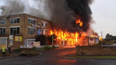 The two storey Marton Hotel and Country Club with flames rising from its windows and fire crews standing by