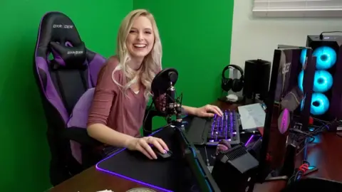 Briana White A young blonde woman sits in a black and purple gaming chair in front of a wall covered in green screen material. On the desk in front of her is a professional streaming set-up - a microphone with pop shield, two monitors, a pair of headphones on a stand and a computer terminal with three fans illuminated by light blue LEDs