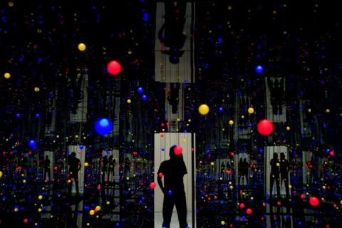 Getty Images A Kusama installation - a dark space with many small coloured dots in the air, and reflections of light doorways with silhouetted figures