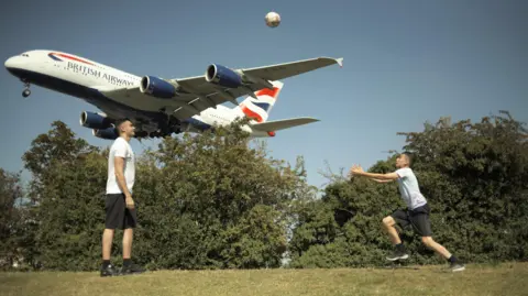  Two boys playing football as a BA plane takes off