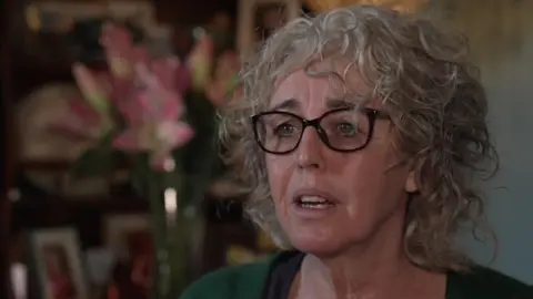 A head and shoulders image of a woman wearing glasses and a green top. She is looking past the camera, seemingly at a television reporter who is out of shot.