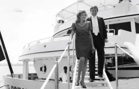 Gambar Getty Foto Hitam dan Putih dari Donald dan Ivana Trump melangkah dari Trump Princess Yacht. Dia mengenakan gaun dan dia mengenakan setelan gelap dengan dasi putih. Kapal pesiar berwarna putih dan laut ada di latar belakang.