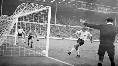 A man in an England top tapping a white football into a goal. Huge crowds watch on. It's a black and white image.