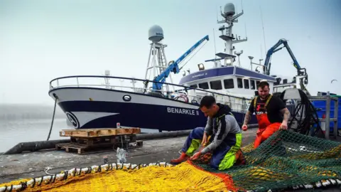 Getty Images A picture of a fishing boat at Peterhead harbour, with two men in high-visibility wading trousers mending nets in the foreground