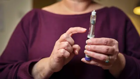 A middle aged woman in a dark purple top is holding a syringe in her left hand. The picture is cropped so you can only see her chest and hands.