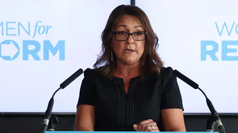 Linden Kemkaran making a speech behind a podium. She is wearing black glasses and a black dress.