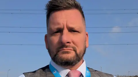 Joel Tetlow has short brown hair and a beard. He is wearing a shirt, waistcoat and pink tie, with the top of a Reform UK lanyard just visible.