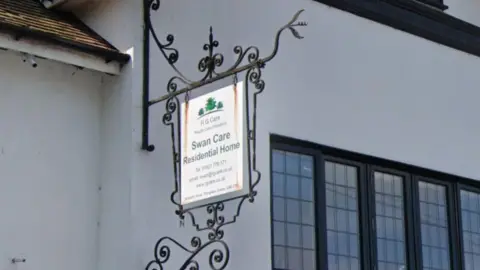 Google A close-up of a white sign hanging outside a white-rendered house, with black framed windows. The sign reads SWAN CARE RESIDENTIAL HOME.