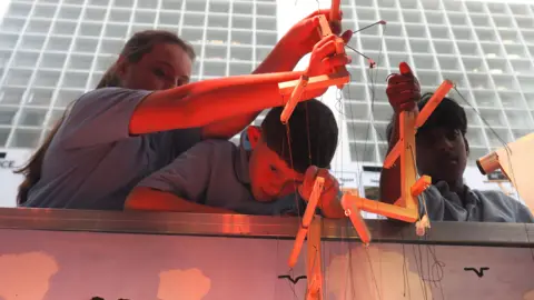 Three schoolchildren controlling puppets (which are out of the picture) using wooden sticks. The children are all wearing grey/light blue short-sleeved shirts. 