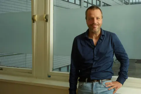 Supplied A man with cropped brown hair and a neat beard smiles as he stands in front of two large windows with cream-coloured frames. He is wearing a dark-blue shirt and light-blue jeans and resting his left hand on his left hip.