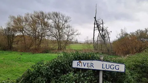 In the foreground is a sign which reads "River Lugg" while in the background is a meadow with trees and bushes.