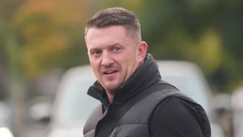 Tommy Robinson looking to the right front of the photo. He is wearing a black top, a black gilet, with a bag strap across one shoulder. He has light stubble.