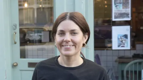 A woman with brown hair that is tied back smiles at the camera. She stands in front of a coffee shop wearing a black long sleeved t-shirt and gold hoop earrings. The shop behind her has a large front window and pale green painted door.