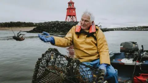 A white-haired man in yellow water proof clothing is sitting on a boat and throwing a lobster back into the sea. He is not far from the shore.