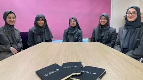 Five schoolgirls wearing black headscarves sitting at a table. 