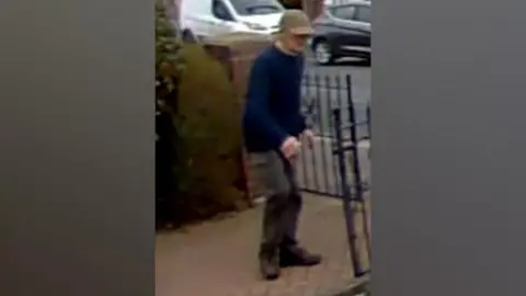 Lancashire Police Brian Fletcher stands on a driveway wearing a blue jumper, tan cap and cargo trousers. His face is slightly hidden by the cap.