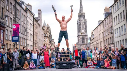 PA Media A street performer holding several sharp objects including a knife and an axe in his hands while he balances on stacked wooden boards in front of a large crowd of peoiple and several old light-stone buildings on the Royal Mile in Edinburgh.