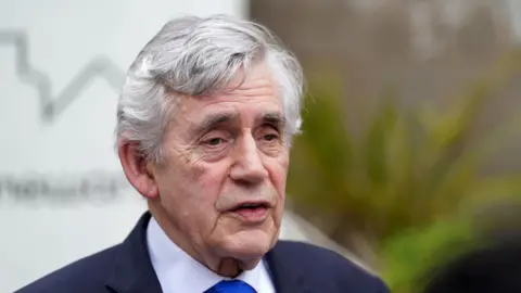 Gordon Brown. He has grey hair and is wearing a dark-coloured suit and royal blue tie. The background is blurred.