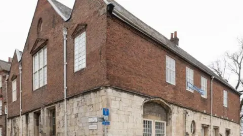 St Anthony's Hall is shown, a large stone and brickwork building. The lower half is a light coloured stone, with the upper half a darker red brick. There are many windows on both the lower and upper halves, with street signs present on the lower. There's also a blue banner hanging from two of the upper windows.