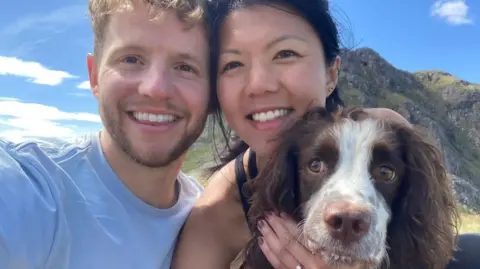 David Parrish and Jade Lau smile at the camera for a selfie with their brown and white spaniel Munro with a mountain in the background on a sunny day. David wears a white t-shirt and Jade wears a black vest