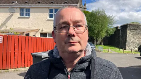 A man with short greying hair stands in front of a house and fence. He is wearing a grey fleece with the collar turned up.