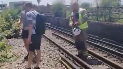 Two train passengers walk alongside train tracks as firefighter guides their direction.
