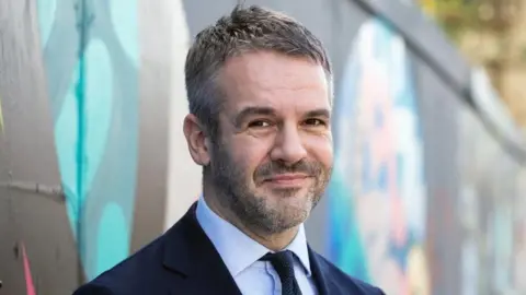 SYMCA South Yorkshire Mayor Oliver Coppard. A smiling man wearing a blue suit and tie and light blue shirt. Behind him is a wall with a colourful mural painted on it.