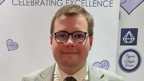 A head and shoulders image of Tom Howard, who is wearing glasses, a grey suit, white shirt and dark grey tie. He is looking straight into the camera. 