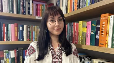 Mariia Kondratiuk is wearing a white dress and is standing in a corner of the library, amongst books in Ukrainian.