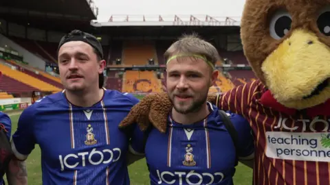 Two men in blue football shirts stand with their arm around each other, in a football stadium. Next to them, a mascot in a chicken costume stands by their side. 